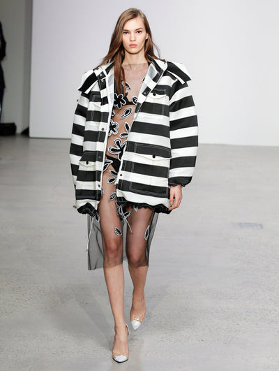 The model walks the runway in a black and white striped oversized coat, layered over a semi-sheer tulle dress with black and white floral embroidery, with part of the audience visible in the background.