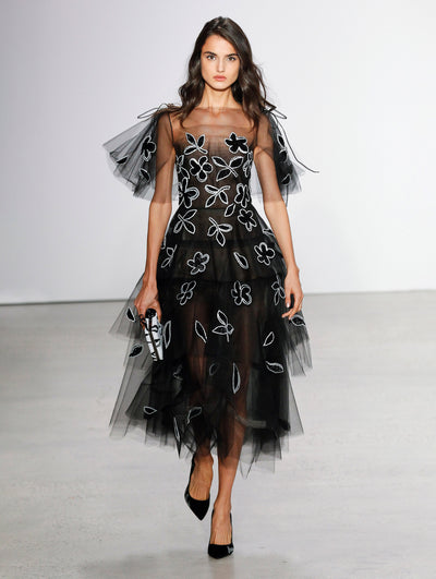 Holding a white and black clutch, the model walks the runway in a semi-sheer tulle tiered cocktail dress with white and black floral embroidery.