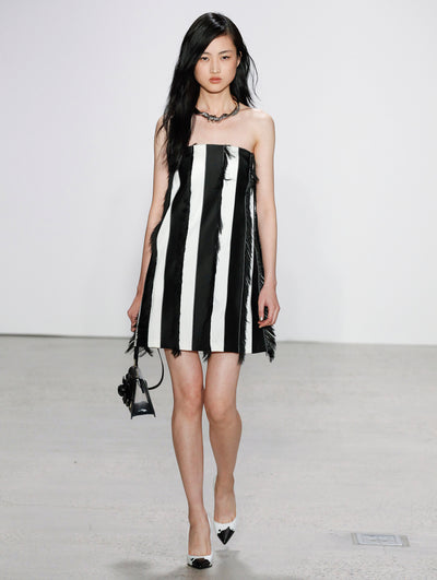 Holding a black gardenia appliqué handbag, the model walks the runway in a strapless black and white vertical striped cocktail dress with fringe, paired with a silver rope-like necklace.