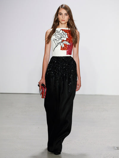 Holding a red and white sequin embroidered clutch, the model walks the runway in a sleeveless white blouse adorned with red brushstroke sequin embroidery and a black flower, paired with black wide-leg trousers featuring degradé sequin embroidery.