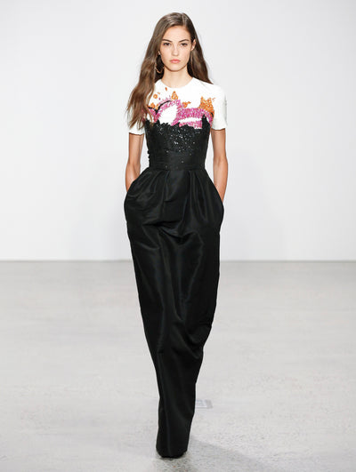 The model walks the runway in a short-sleeve column gown. The gown has a white bodice decorated with orange and pink sequin floral embroidery, transitioning into a black skirt embellished with black sequin embroidery.