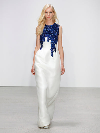 The model walks the runway in a sleeveless white column gown, with the bodice adorned in cascading blue sequin embroidery.