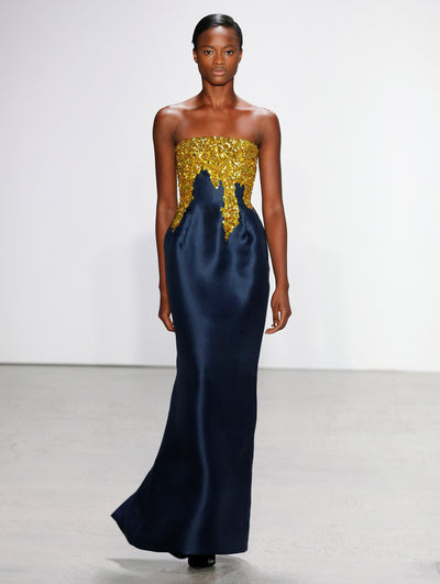 The model walks the runway in a strapless navy column gown, with the bodice adorned in cascading gold sequin embroidery.