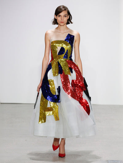 The model walks the runway in an illusion-neck semi-sheer white cocktail dress, decorated with large-scale sequin-embroidered letters spelling 'Oscar de la Renta' in shades of blue, gold and red.