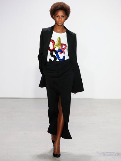 A model walks the runway in a black long-sleeve blazer, layered over a black maxi skirt, and a white tee adorned with large-scale sequin-embroidered letters spelling 'Oscar' in shades of blue, gold and red.
