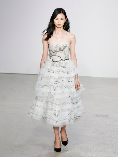 The model walks the runway in a strapless white tulle tiered cocktail dress printed with the Oscar de la Renta logo in varying sizes.