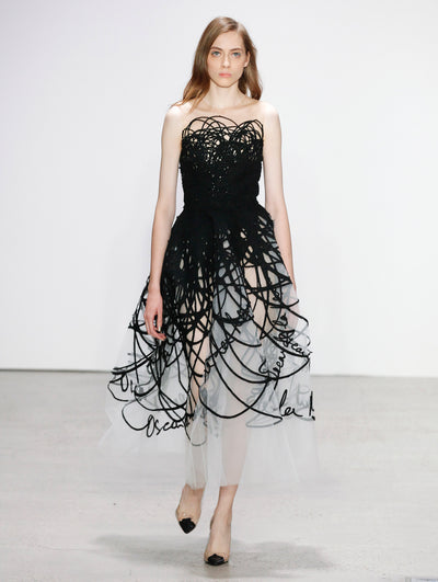 The model walks the runway in a white semi-sheer strapless tulle cocktail dress featuring all-over black brush strokes that converge into a fully black bodice, with the Oscar de la Renta logo delicately spaced throughout.