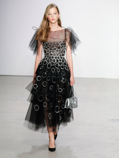 Holding a silver gardenia appliqué handbag, the model walks the runway in a semi-sheer tulle tiered cocktail dress with crystal embroidery forming circles concentrated at the bodice.