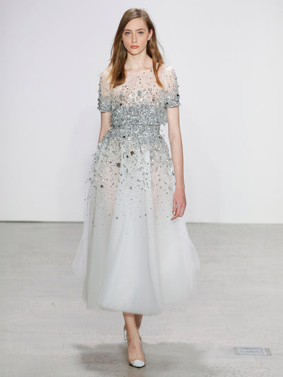 The model walks the runway in a white tulle short-sleeve cocktail dress with silver sequin embroidery, concentrated at the bodice and fading into a delicate scatter toward the skirt.
