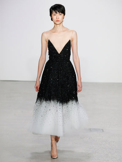 The model walks the runway in an illusion-neck tulle cocktail dress, the bodice is black, and bodice is adorned with cascading tonal sequin embroidery. The skirt transitions to white with black sequin embroidery. 
