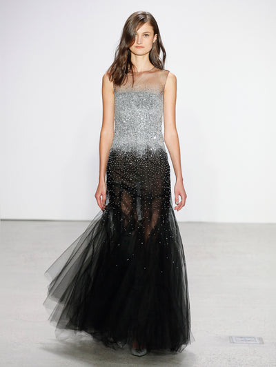 The model walks the runway in a semi-sheer illusion-neck tulle black evening gown adorned with silver crystal embroidery concentrated at the bodice, fading into a delicate scatter toward the skirt.