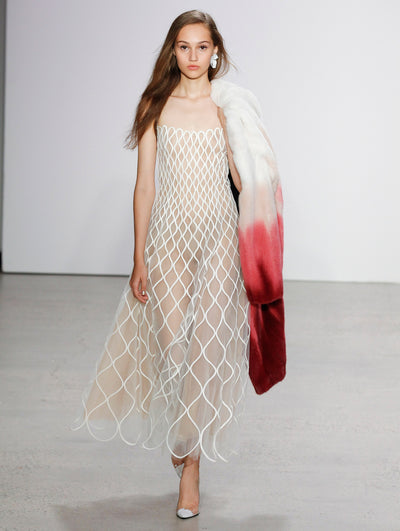 The model walks the runway in a strapless semi-sheer white tulle cocktail dress, adorned with sinuous white embroidery, while holding an ombré fur coat where shades of bright white transition to deep red.