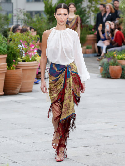 The model walks the runway in a white long-sleeve one-shoulder blouse and silk wrap skirt with a multi-color tapestry design and fringe trim. Behind her, the blurred background reveals the audience and potted plants.