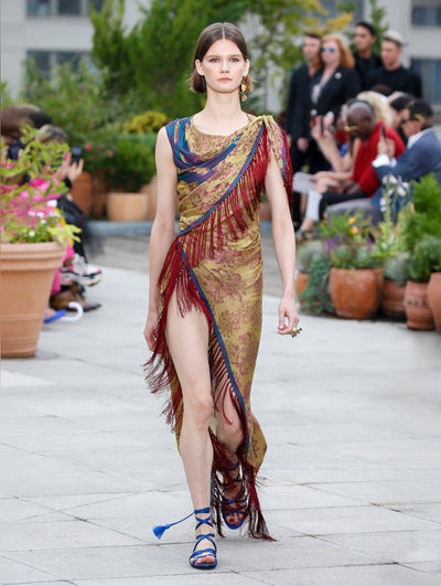 The model walks the runway in a silk wrap dress with a multi-colored tapestry design and fringe trim. Behind her, the blurred background reveals the audience and potted plants.
