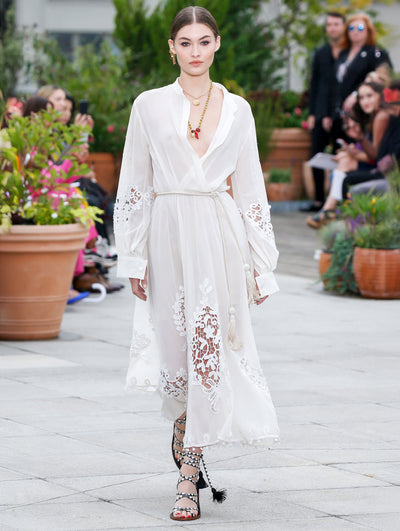 The model walks the runway in a white long-sleeve V-neck dress with lace insets and pompom trim. Behind her, the blurred background reveals the audience and potted plants.