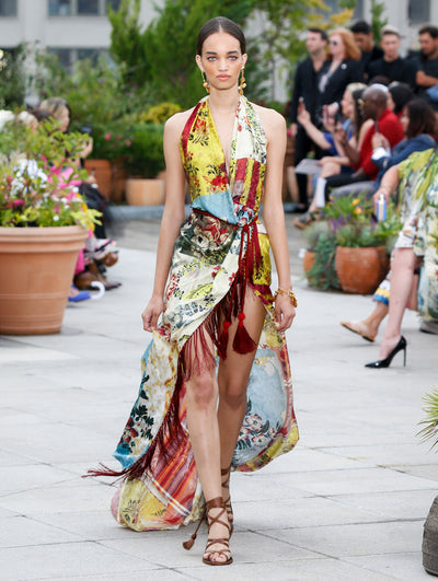 The model walks the runway in a sleeveless halter-neck silk dress with a multi-colored tapestry design and fringe trim. Behind her, the blurred background reveals the audience and potted plants.