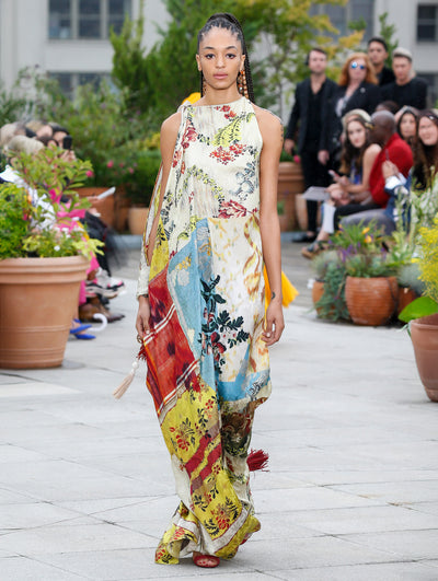 The model walks the runway in a sleeveless silk dress with a multi-colored tapestry design. Behind her, the blurred background reveals the audience and potted plants.