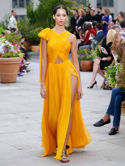 The model walks the runway in an orange sleeveless bow-detailed evening gown with a front slit. Behind her, the blurred background reveals the audience and potted plants.
