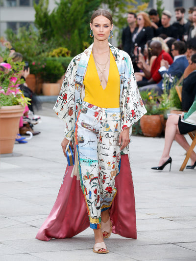 The model walks the runway in an open-front silk coat printed with a multi-color map design, layered over a yellow V-neck blouse and silk trousers printed with the same map design. Behind her, the blurred background reveals the audience and potted plants.