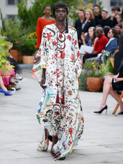 The model walks the runway in a V-neck silk caftan printed with a multi-colored floral and map design. Behind her, the blurred background reveals the audience and potted plants.