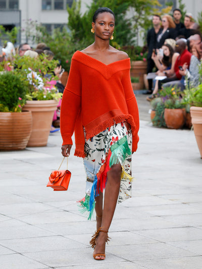 Holding a red gardenia appliqué handbag, the model walks the runway in a red off-shoulder long-sleeve knit pullover with fringe trim, paired with a wrap silk skirt printed with a floral design and multi-colored fringe trim. Behind her, the blurred background reveals the audience and potted plants.