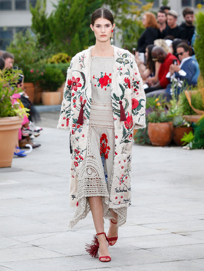 The model walks the runway in a beige floral-embroidered coat, layered over a beige crochet dress with red floral embroidery. Behind her, the blurred background reveals the audience and potted plants.