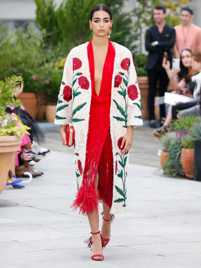 Holding a red clutch, the model walks the runway in a beige floral-embroidered coat, layered over a red V-neck cocktail dress with sequin embroidery and feather trim. Behind her, the blurred background reveals the audience and potted plants.