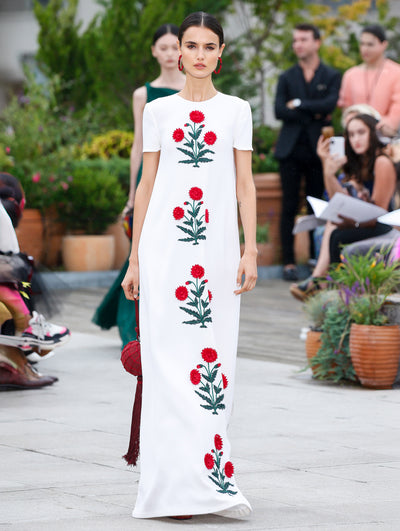 The model walks the runway in white short-sleeve column dress with multi-colored threadwork embroidered floras at the front. Behind her, the blurred background reveals the audience and potted plants.
