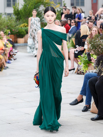 Holding a bird-shaped handbag, the model walks the runway in green off-shoulder evening gown with an open bodice. Behind her, the blurred background reveals the audience and potted plants.