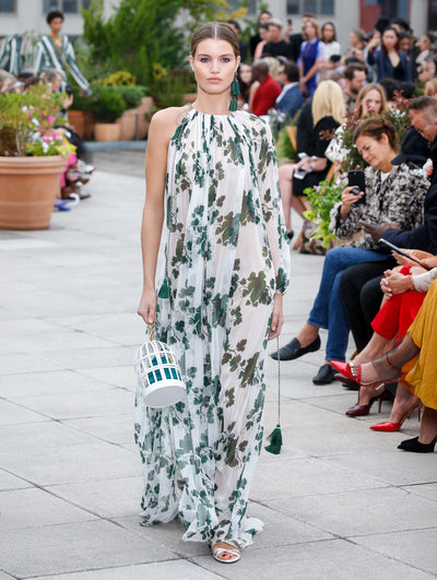 Holding a birdcage-shaped handbag, the model walks the runway in white one-shoulder long-sleeve silk caftan printed with green leaves. Behind her, the blurred background reveals the audience and potted plants.