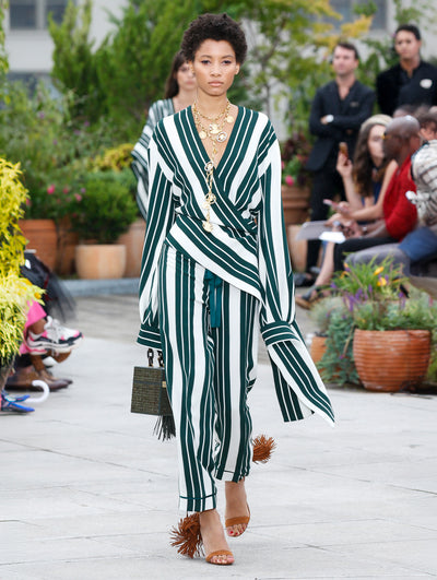 Holding a green woven cube-like handbag, the model walks the runway in a white and green striped long-sleeve blouse with an asymmetrical hemline and coordinating trousers with the same striped design. Behind her, the blurred background reveals the audience and potted plants.