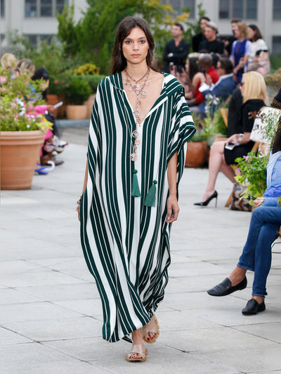 The model walks the runway in a white and green striped V-neck silk caftan. Behind her, the blurred background reveals the audience and potted plants.