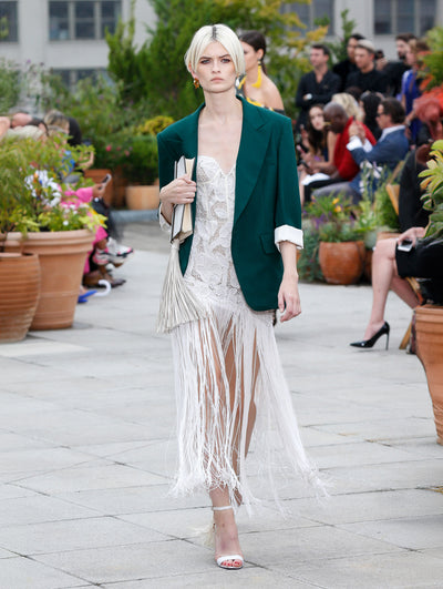 Holding an ivory clutch, the model walks the runway in a green single-breasted blazer, layered over an ivory sweetheart threadwork embroidered cocktail dress with fringe trim. Behind her, the blurred background reveals the audience and potted plants.