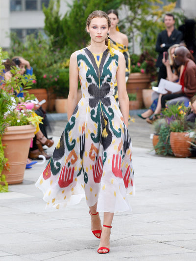 The model walks the runway in a white 
 sleeveless fil coupé cocktail dress printed with a multi-colored abstract tulip design. Behind her, the blurred background reveals the audience and potted plants.