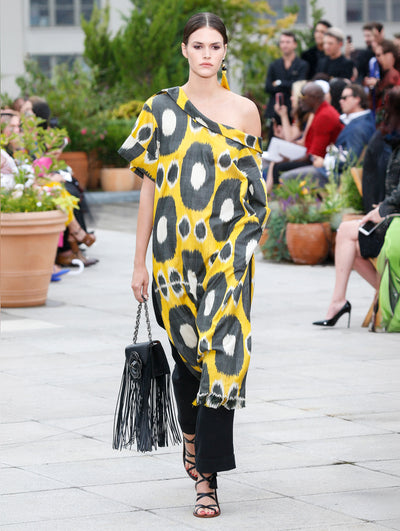 Holding a black gardenia appliqué handbag with fringe, the model walks the runway in a one-shoulder short-sleeve tunic with a large-scale polka dot design, layered over black trousers. Behind her, the blurred background reveals the audience and potted plants.