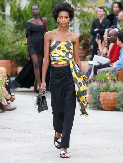 Holding a black gardenia appliqué handbag with fringe, the model walks the runway in a strapless blouse with a large-scale polka dot design and drape detailing, paired with black trousers. Behind her, the blurred background reveals the audience and potted plants.