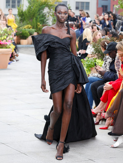 The model walks the runway in a black off-shoulder bow-detailed cocktail dress with a floor-sweeping asymmetrical hemline. Behind her, the blurred background reveals the audience and potted plants.