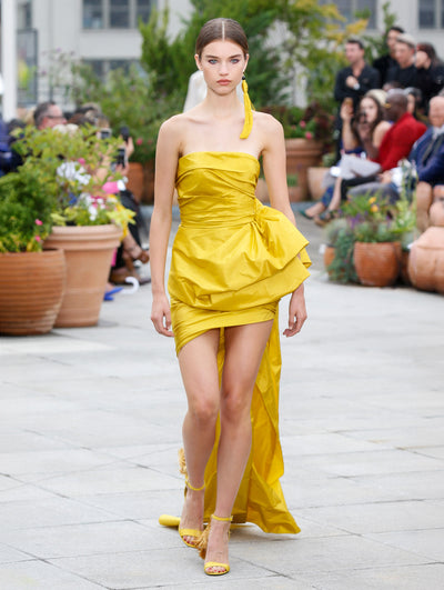 The model walks the runway in a yellow strapless drape-detailed cocktail dress with a floor-sweeping asymmetrical hemline. Behind her, the blurred background reveals the audience and potted plants.