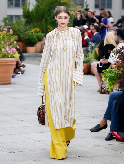 Holding a leather birdcage-shaped handbag, the model walks the runway in a white and yellow stripped long-sleeve bow-detailed tunic, layered over yellow trousers. Behind her, the blurred background reveals the audience and potted plants.