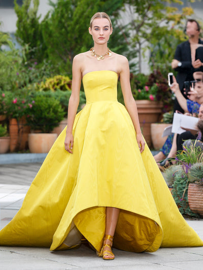 The model walks the runway in a yellow strapless high-low ball gown with short train. Behind her, the blurred background reveals the audience and potted plants.