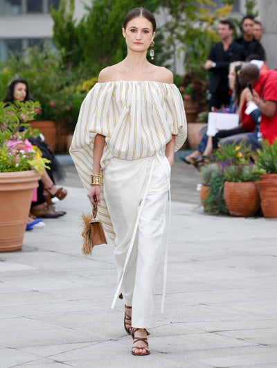 Holding a brown gardenia appliqué handbag with fringe detailing, the model walks the runway in an off-shoulder white and yellow striped blouse, paired with white trousers. Behind her, the blurred background reveals the audience and potted plants.