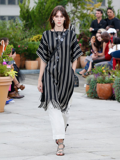 Wearing a black crossbody bag, the model walks the runway in a black and white striped knit tunic with fringe trim, layered over white trousers. Behind her, the blurred background reveals the audience and potted plants.
