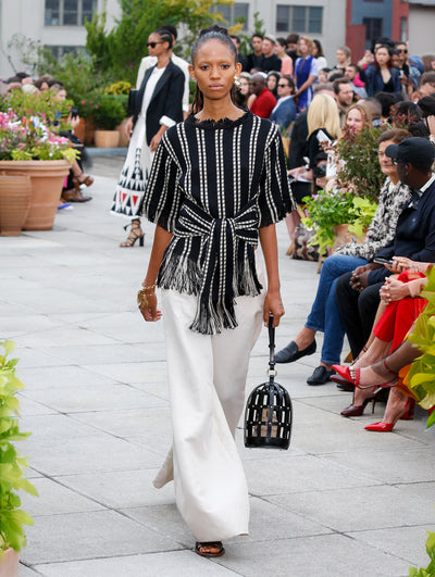 Holding a leather birdcage-shaped handbag, the model walks the runway in a black and white striped knit blouse with fringe trim, paired with white trousers. Behind her, the blurred background reveals the audience and potted plants.