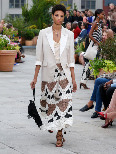 Holding a black bird-shaped handbag, the model walks the runway in a cream single-breasted blazer with cuffed sleeves, layered over a satin blouse and a black, white and brown crocheted skirt with a geometric design. Behind her, the blurred background reveals the audience and potted plants.