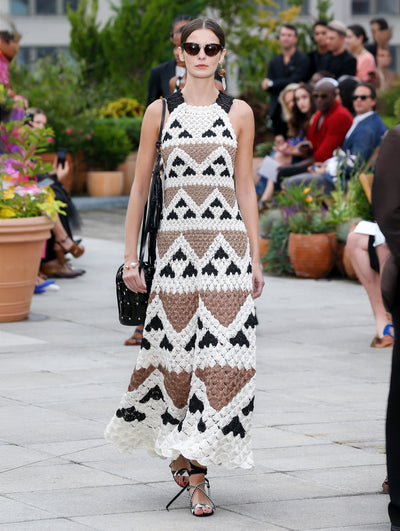 Wearing a birdcage-shaped black shoulder bag, the model walks the runway in black, white and brown crocheted maxi dress with a geometric design. Behind her, the blurred background reveals the audience and potted plants.