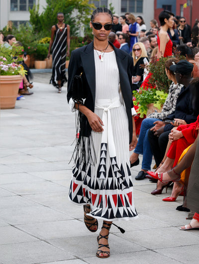 Wearing black sunglasses and holding a black clutch, the model walks the runway in navy blazer with cuffed sleeved, layered over a white knit dress with a multi-colored geometric design concentrated on the skirt. Behind her, the blurred background reveals the audience and potted plants.