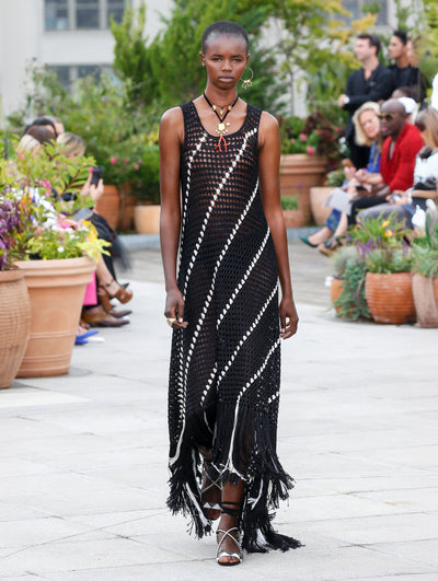 The model walks the runway in a semi-sheer black and white striped crochet maxi dress with fringe trim. Behind her, the blurred background reveals the audience and potted plants.