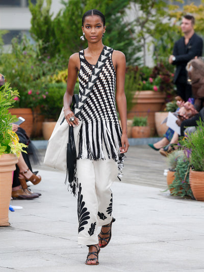 Wearing a crossbody white duffle bag, the model walks the runway in a black and white striped crochet top with fringe trim, paired with white trousers embroidered with black vines. Behind her, the blurred background reveals the audience and potted plants.