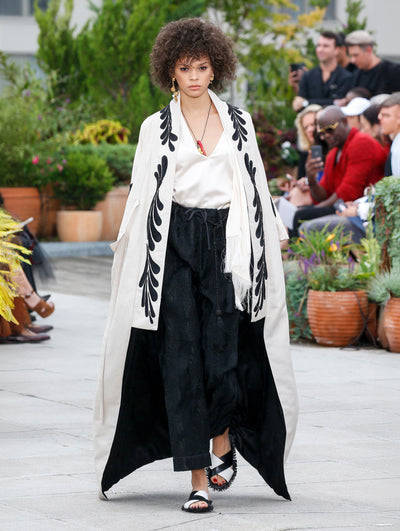 The model walks the runway in a white floor-length kimono-inspired coat with black threadwork embroidered vines, layered over a white V-neck blouse and black trousers. Behind her, the blurred background reveals the audience and potted plants.