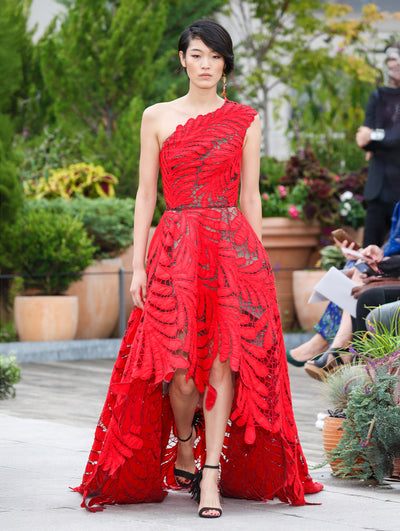 The model walks the runway in a red one-shoulder high-low evening gown with a threadwork cutout fern design. Behind her, the blurred background reveals the audience and potted plants.